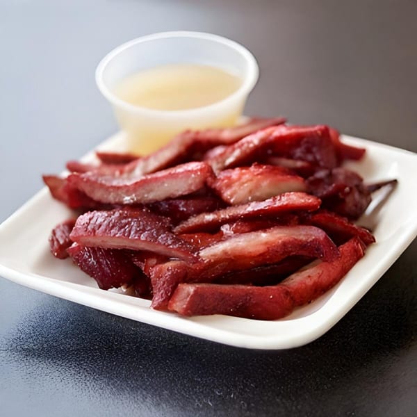 A white plate of sliced red char siu pork with a small cup of sauce.