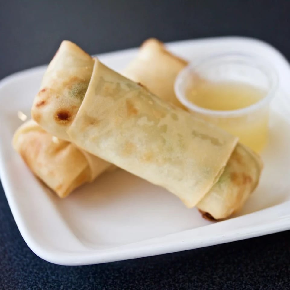 Two crispy egg rolls with a side of dipping sauce on a white plate from Lotus Xpress.